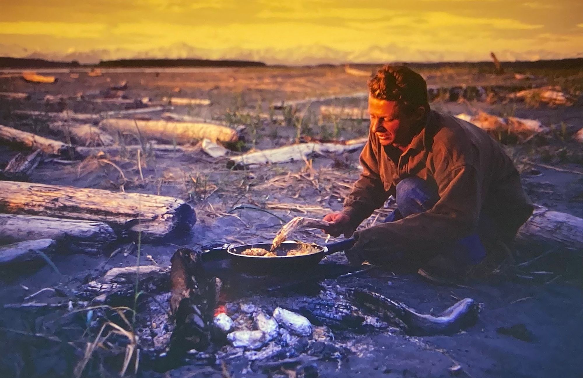 Patrick Trefz cooking by a campfire as the sun sets in Alaska